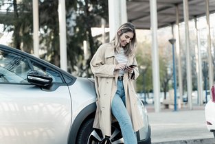 Kobieta w beżowym płaszczu pisząca wiadomość na telefonie jest oparta o srebrny samochód 